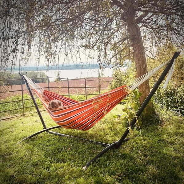 A hammock tied between two trees, overlooking a grassy area with a view of a body of water and distant landscape. - The Riverside Retreat (GLOUCESTERSHIRE)
