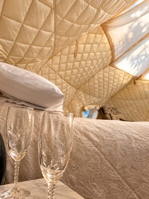 A tented accommodation with a quilted interior, a white pillow, and two empty wine glasses on a table. - The Riverside Retreat (GLOUCESTERSHIRE)