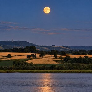 Un paisaje con un cuerpo de agua reflejando la luna, colinas ondulantes y una torre distante.