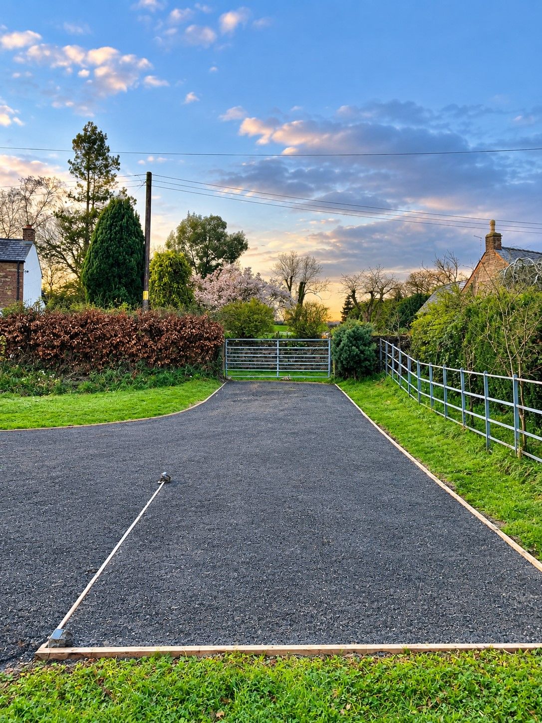 A paved driveway leading to a gated entrance with a metal gate, surrounded by greenery and a clear sky.