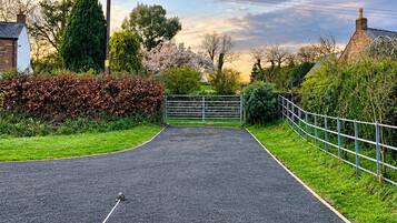 A paved driveway leading to a gated entrance with a metal gate, surrounded by greenery and a clear sky.