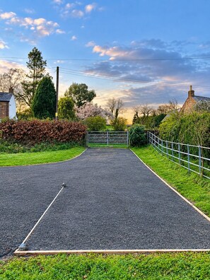 A paved driveway leading to a gated entrance with a metal gate, surrounded by greenery and a clear sky. - The Riverside Retreat (GLOUCESTERSHIRE)