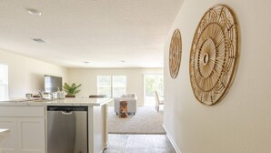 A modern kitchen with white cabinetry, a dishwasher, and a dining area with a white sofa and a small wooden stool. - Beautiful New 4-BR Home Near All Major Attractions (Palm Coast)
