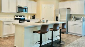 A modern kitchen with white cabinets, a central island with bar stools, stainless steel appliances, and a built-in microwave. - Beautiful New 4-BR Home Near All Major Attractions (Palm Coast)