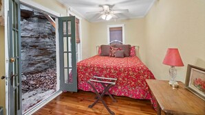 A bedroom with a bed, a nightstand, a lamp, a door with a stone wall, and a ceiling fan.