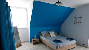 Une chambre avec un plafond en pente, un lit recouvert d’une courtepointe bleue, une table de chevet avec une lampe et une fenêtre avec des rideaux.