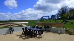 Une terrasse extérieure avec une table et des chaises, un barbecue et, au fond, un ensemble de jeux pour enfants avec une balançoire.
