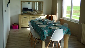 Un coin repas avec une table dressée pour six personnes, des chaises, un buffet en bois et une fenêtre donnant sur un paysage verdoyant.
