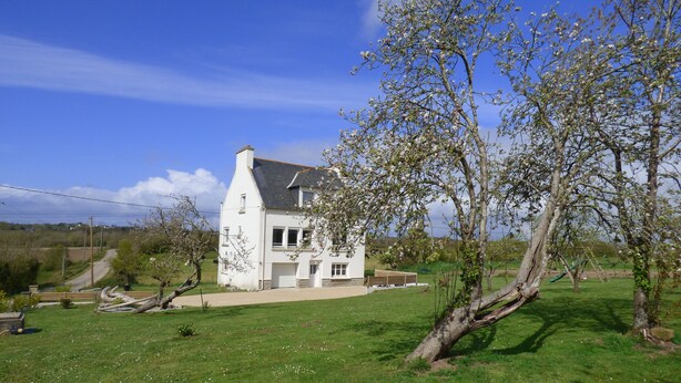 Une maison blanche à deux étages, avec un toit en pente, entourée d’une pelouse verdoyante et d’arbres.
