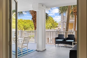 Un espacio al aire libre techado con barandilla blanca, una mesa con sillas y vistas a una zona residencial con árboles y un edificio.