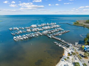 An aerial view of a marina with numerous docks and boats, surrounded by clear blue water and a distant shoreline.