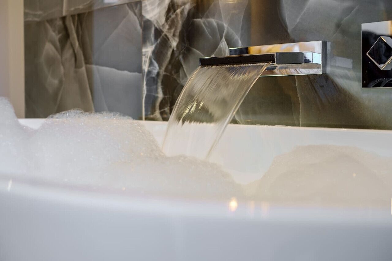 A modern bathroom with a chrome faucet, a marble wall, and a bathtub filled with bubbles.