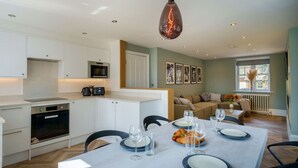 Eine moderne Küche mit weißen Schränken, ein Essbereich mit Tisch für vier Personen und ein Wohnbereich mit Sofa und Fenster.