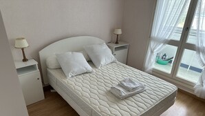 A neatly made bed with white bedding, two bedside tables with lamps, and a view of the outside through a window with sheer curtains.