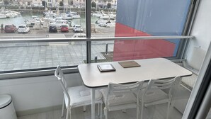 A room with a view of a marina, a white table with chairs, a white trash bin, and a window with a view of parked cars and boats.
