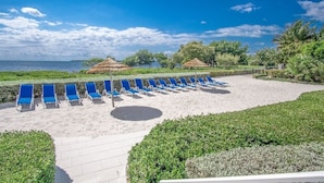 En strand med rader av blå solstolar, ett sandområde och byggnader med halmtak.