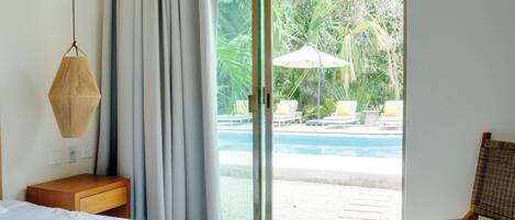 A hotel room with a bed, a wooden nightstand, a wicker chair, and a view of a pool and outdoor seating area.