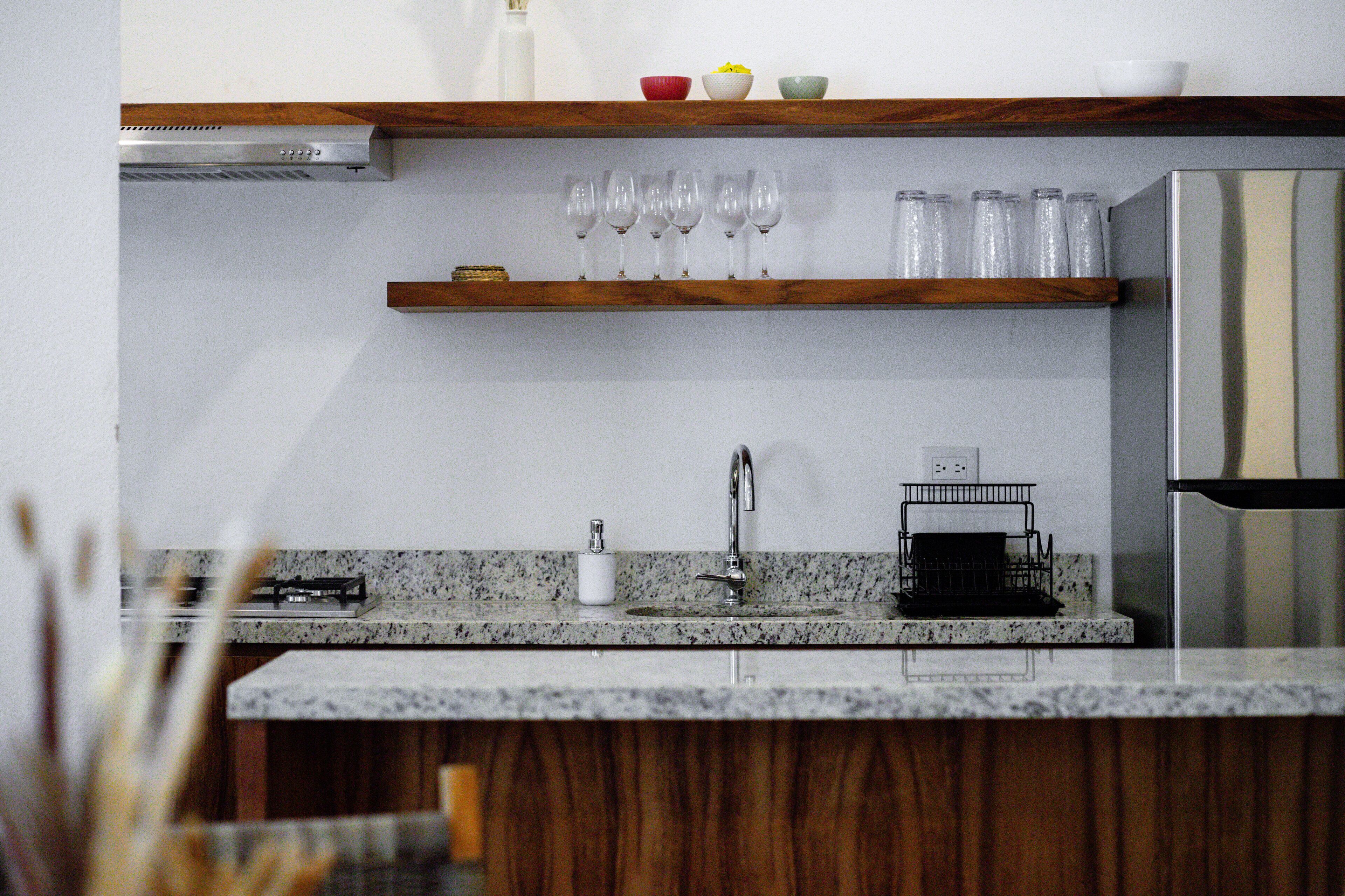 Una cocina con encimeras de granito, un refrigerador de acero inoxidable y un estante de madera sobre el lavabo con vasos y cuencos.