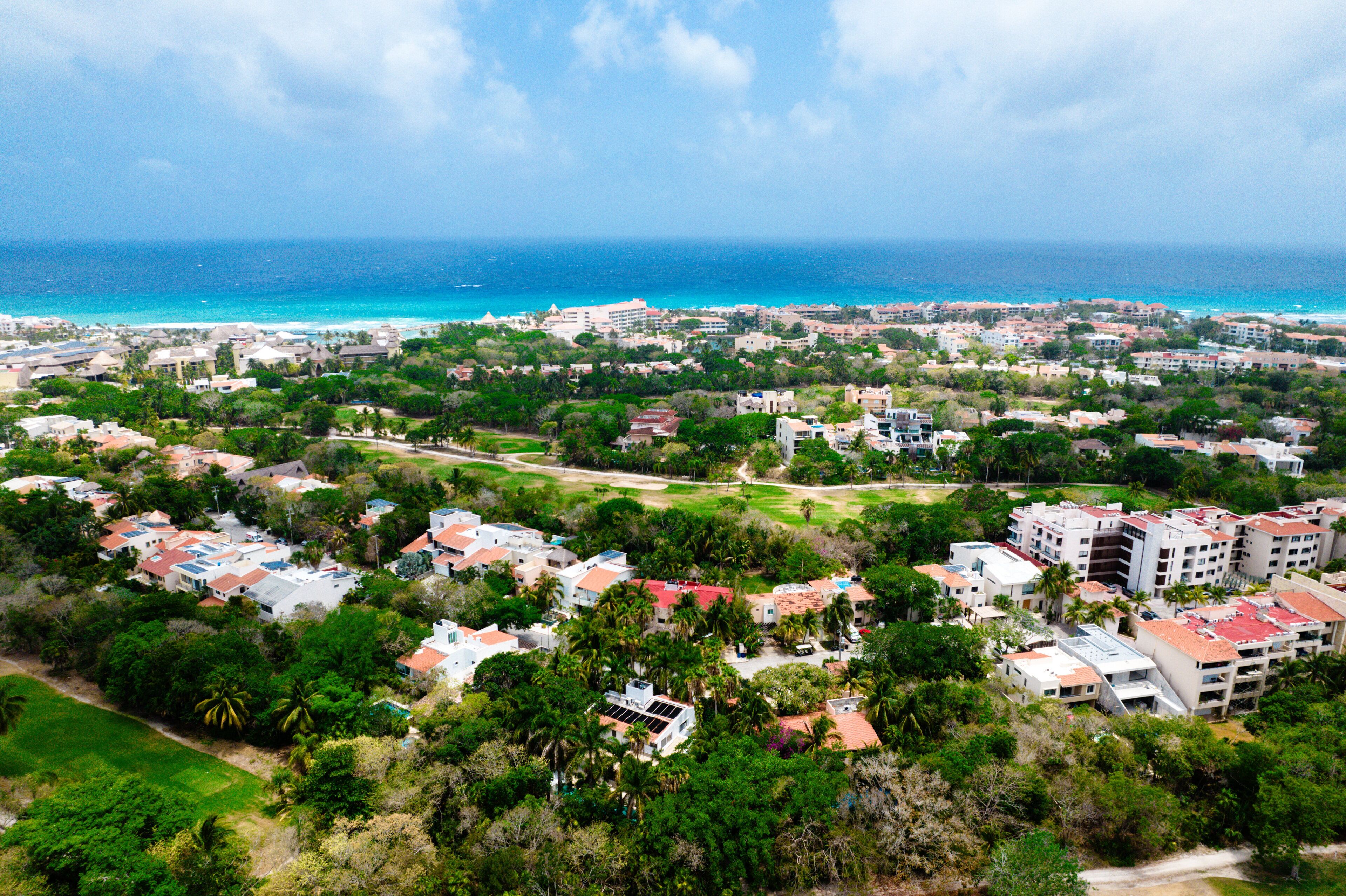 An aerial view of a coastal residential area with houses, green spaces, and a view of the ocean.