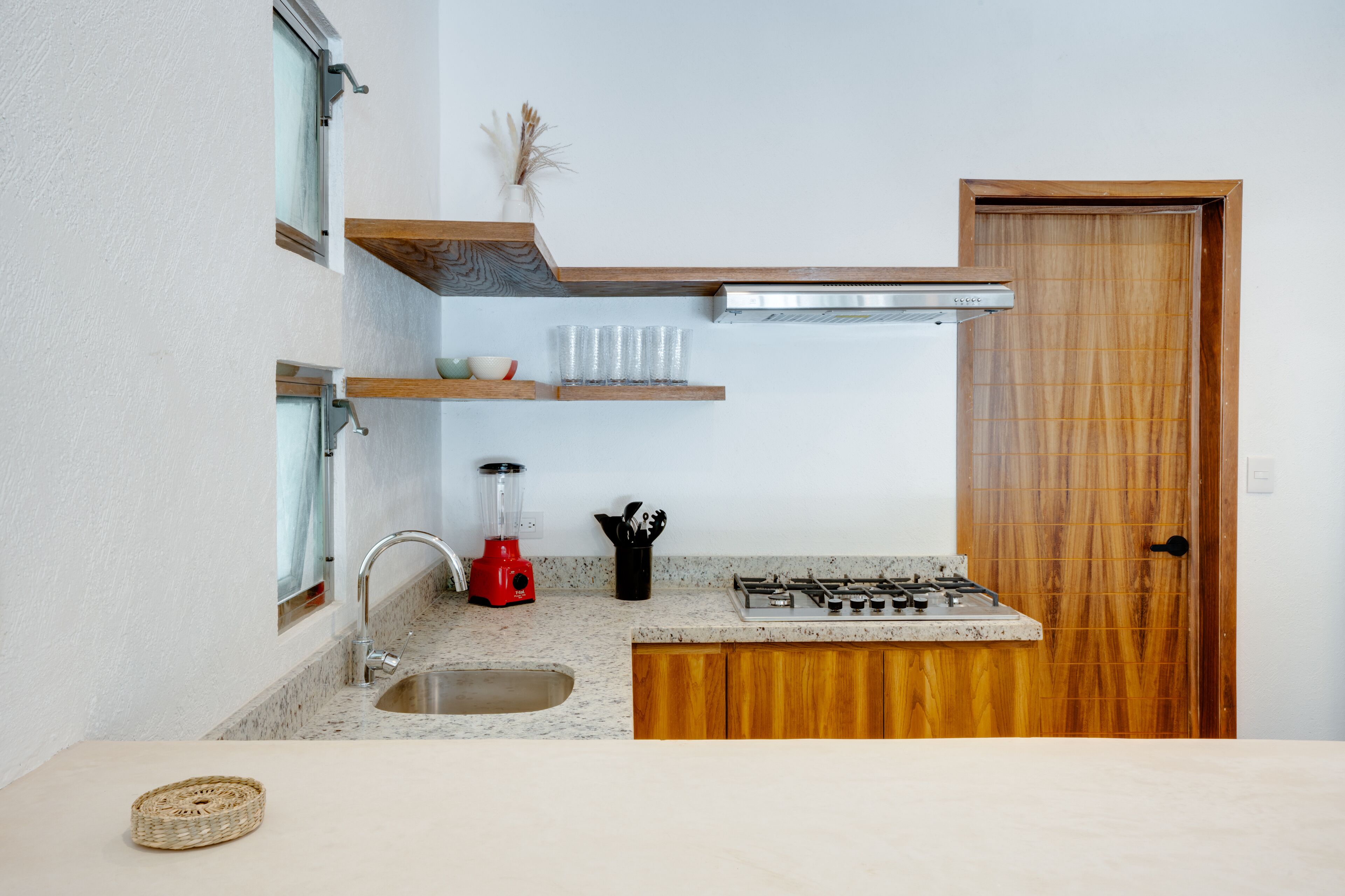 A compact kitchen with a granite countertop, a stainless steel sink, a red blender, a gas stove, and wooden shelves.