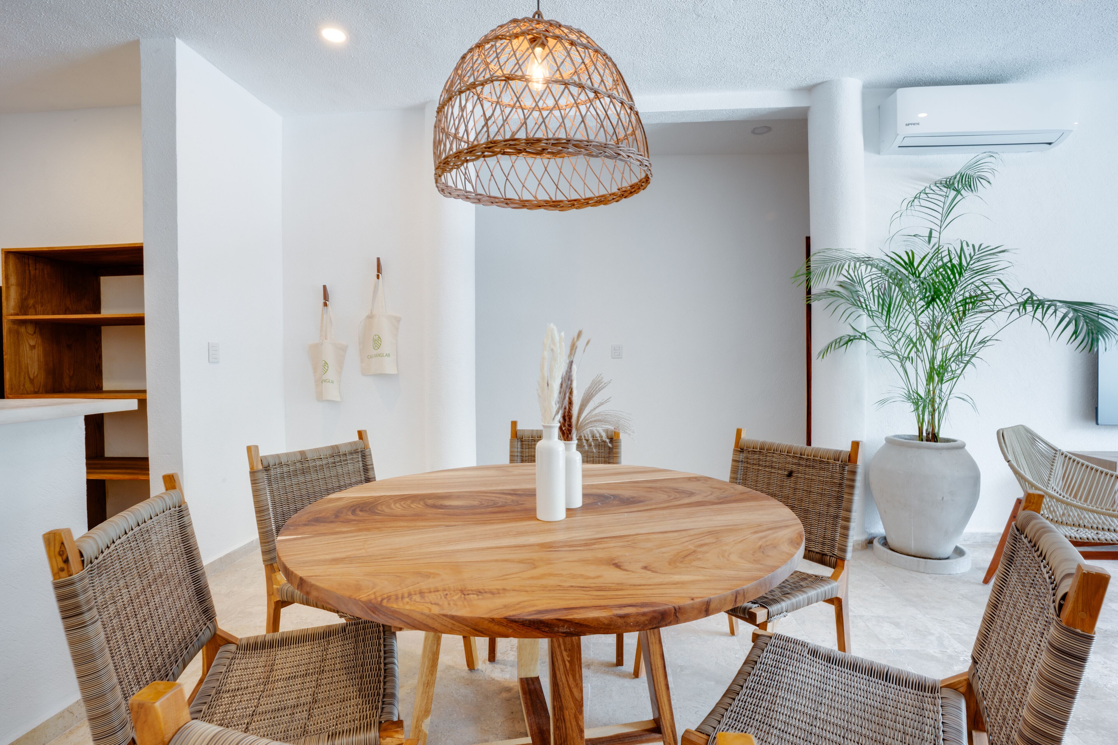 Una mesa de madera redonda rodeada de sillas de mimbre, una lámpara colgante tejida y una planta grande en maceta.