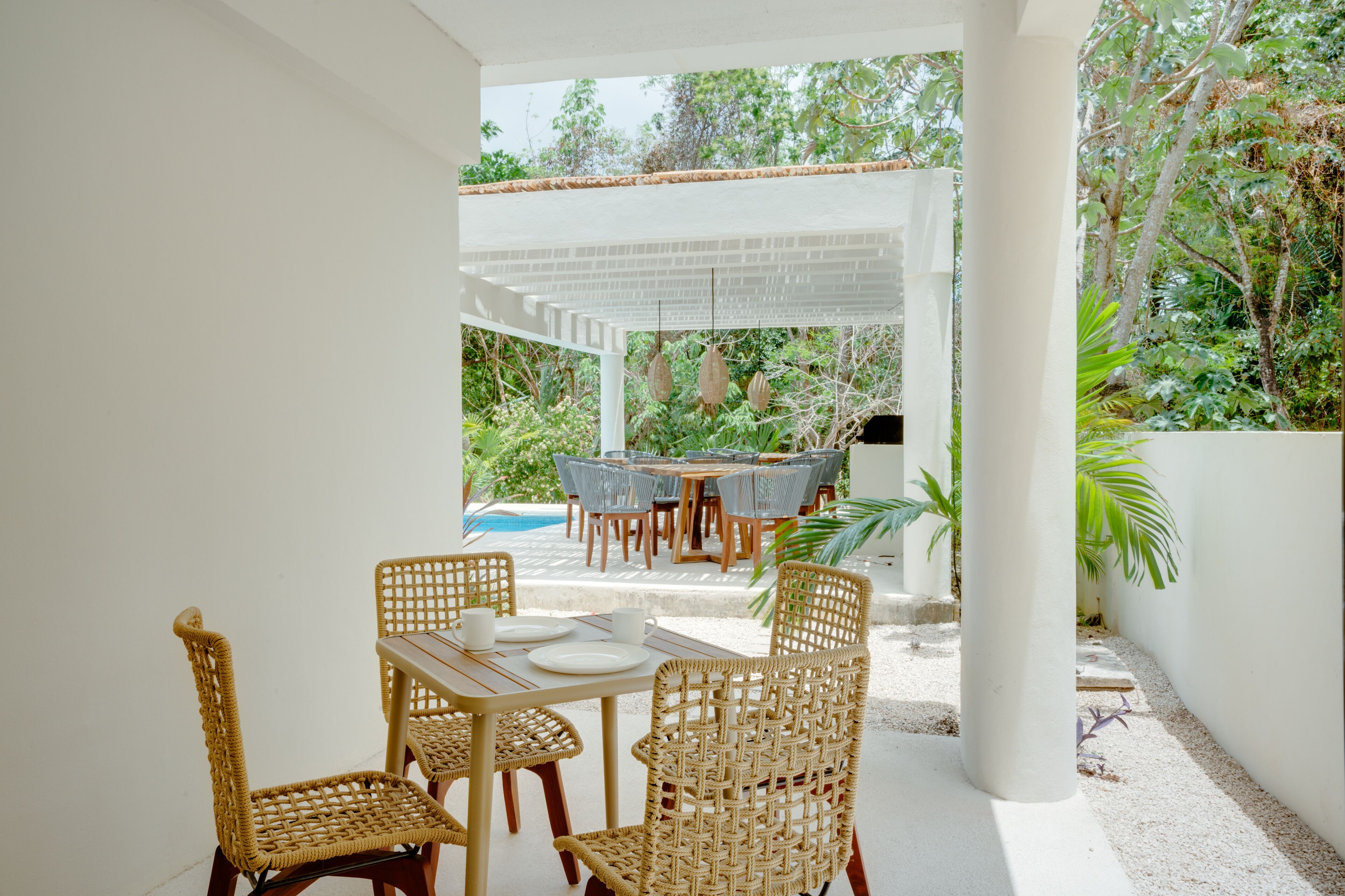 Un patio exterior con una pérgola blanca, sillas de mimbre, una mesa de madera y una piscina al fondo.