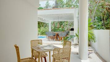 Un patio exterior con una pérgola blanca, sillas de mimbre, una mesa de madera y una piscina al fondo.