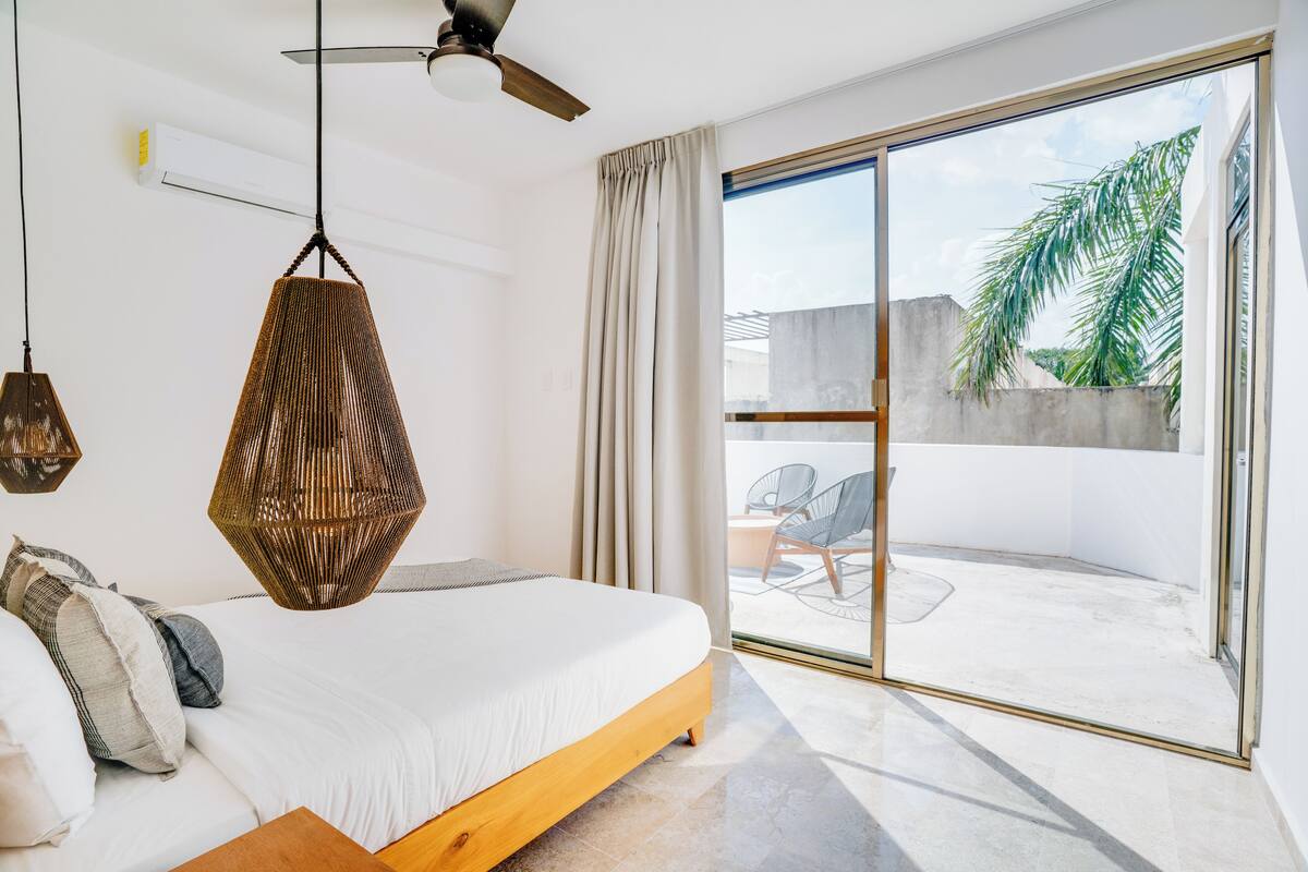 Un dormitorio moderno con cama, lámpara colgante, ventilador de techo y vistas a un patio exterior con sillas.