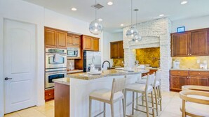 A modern kitchen with wooden cabinets, a central island with bar stools, and a stone accent wall. - Arcade Blast/Pool/Spa/100k Game Room (Indio)