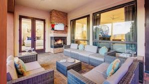 A cozy outdoor seating area with wicker furniture, a fireplace, and large windows offering a view of the outdoors. - Arcade Blast/Pool/Spa/100k Game Room (Indio)
