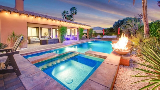 A swimming pool with a fire feature, a covered patio with outdoor furniture, and a house with a tiled roof and large windows. - Arcade Blast/Pool/Spa/100k Game Room (Indio)