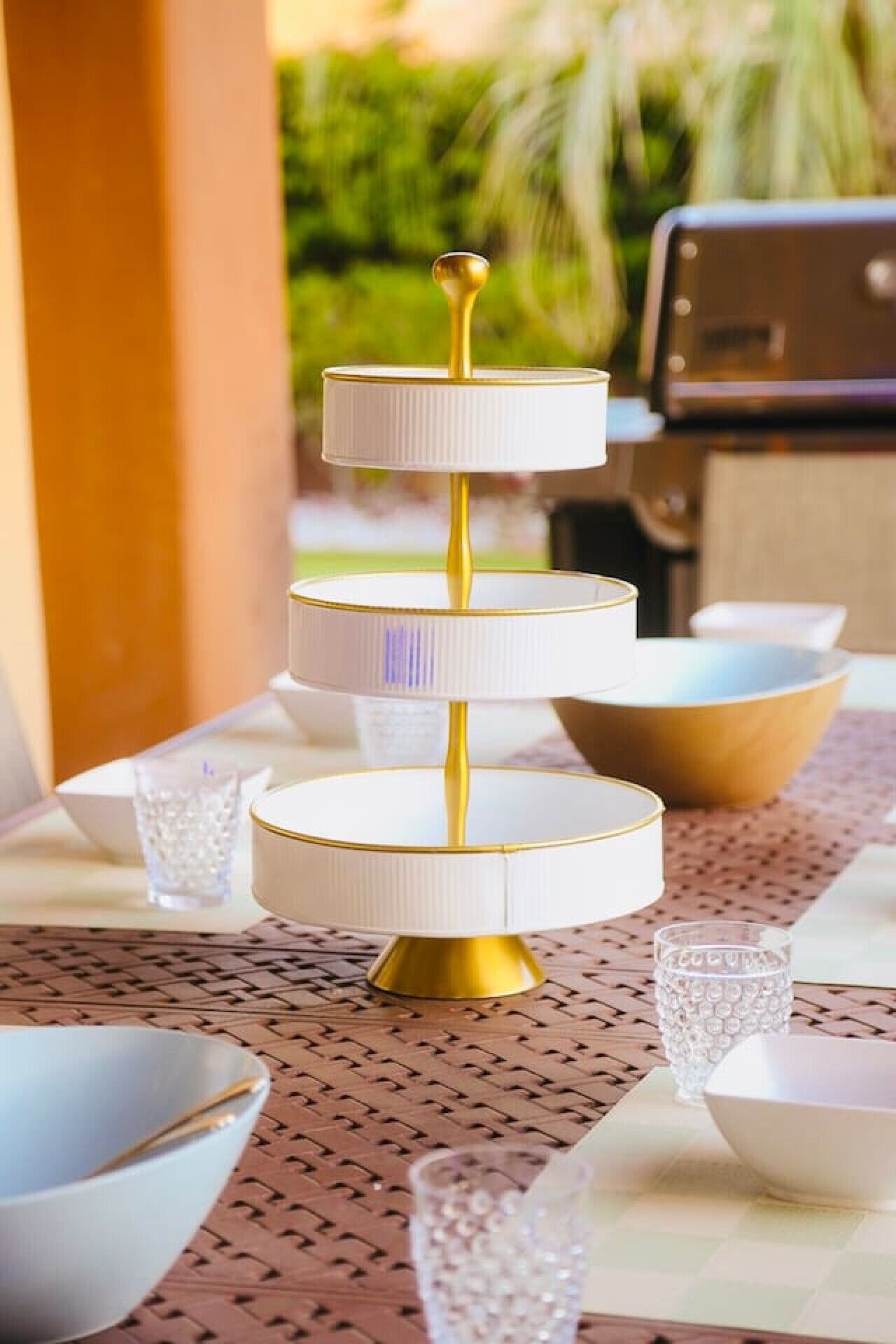 Un plateau de service à trois niveaux, avec une base dorée et des assiettes blanches, posé sur une table recouverte d’un set de table en fibres tressées.