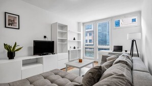 Un salon moderne avec un canapé gris, un téléviseur à écran plat, des étagères blanches, une petite table et un lampadaire.