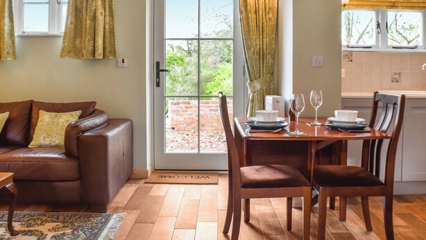 Una sala acogedora con un sofá de cuero marrón, una mesa de comedor de madera para dos personas y vistas a un muro de ladrillo y vegetación exterior.