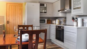 Cocina compacta con armarios blancos, horno empotrado, microondas y refrigerador. Hay una mesa de comedor de madera con sillas y una copa de vino encima.