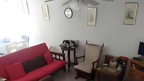 Ein Wohnzimmer mit einem roten Sofa, zwei braunen Sesseln, einem Holztisch und einem kleinen Tisch mit einem Teddybären. Eine Deckenventilator, eine Uhr und ein gerahmtes Bild sind an der Wand angebracht.