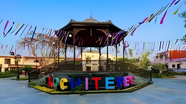Eine Laube mit bunten Buchstaben, die „MOTELERIA“ auf ihrem Fuß zeigen, umgeben von blauem Himmel und ruhigem Gewässer.