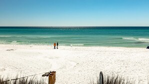 En strand med vit sand, klart blått vatten och några få personer som promenerar.