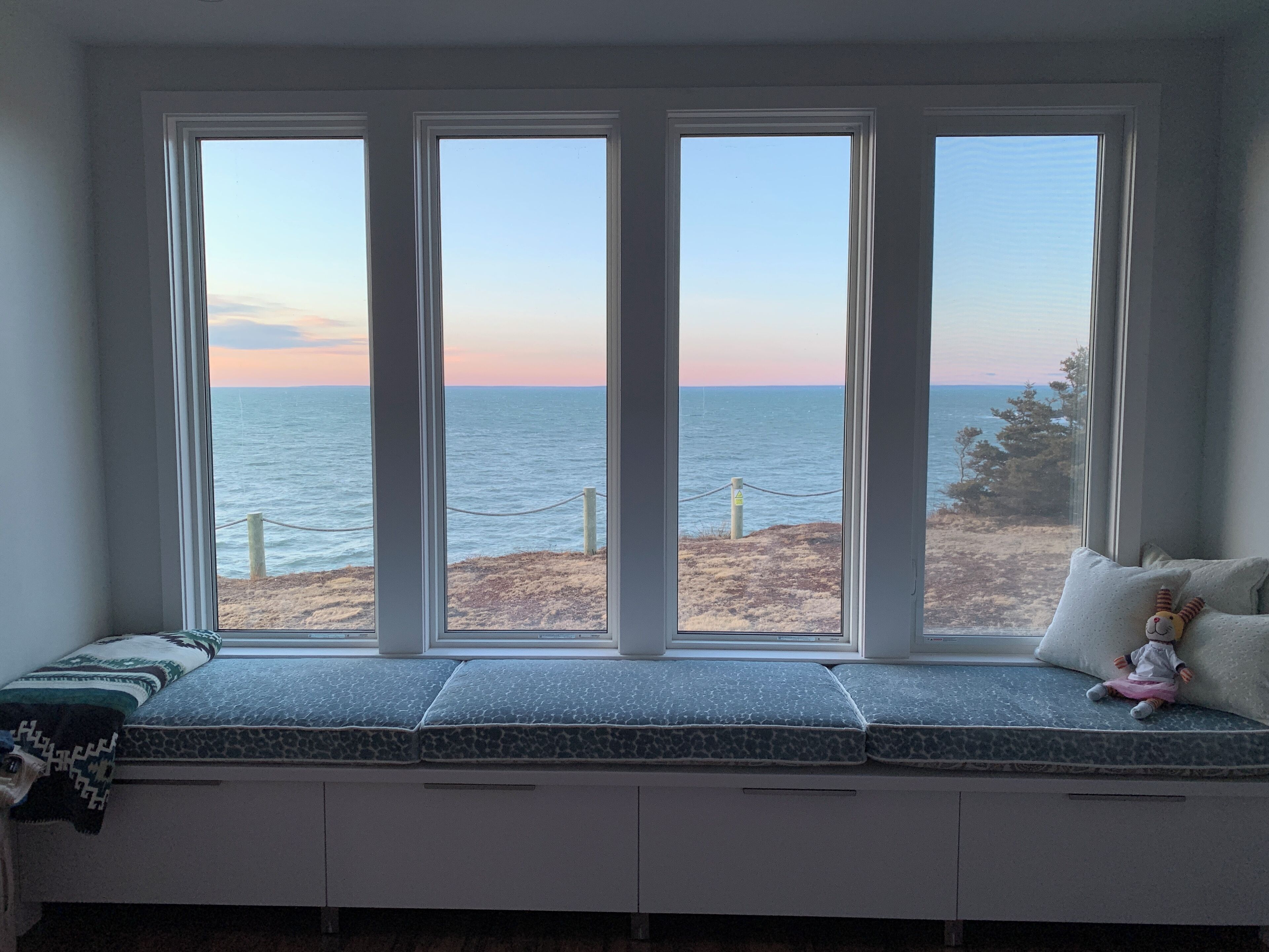 A room with a large window overlooking a beach, a bench with cushions, and a small stuffed toy on the bench.