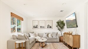 A modern living room with a sectional sofa, a small round table, and a wooden cabinet. There are three framed pictures on the wall and a large window with a view of trees.