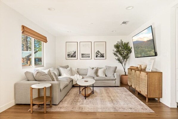 Un salon moderne avec un canapé d’angle, une petite table ronde et un meuble en bois. Trois tableaux encadrés ornent le mur, et une grande fenêtre offre une vue sur les arbres.