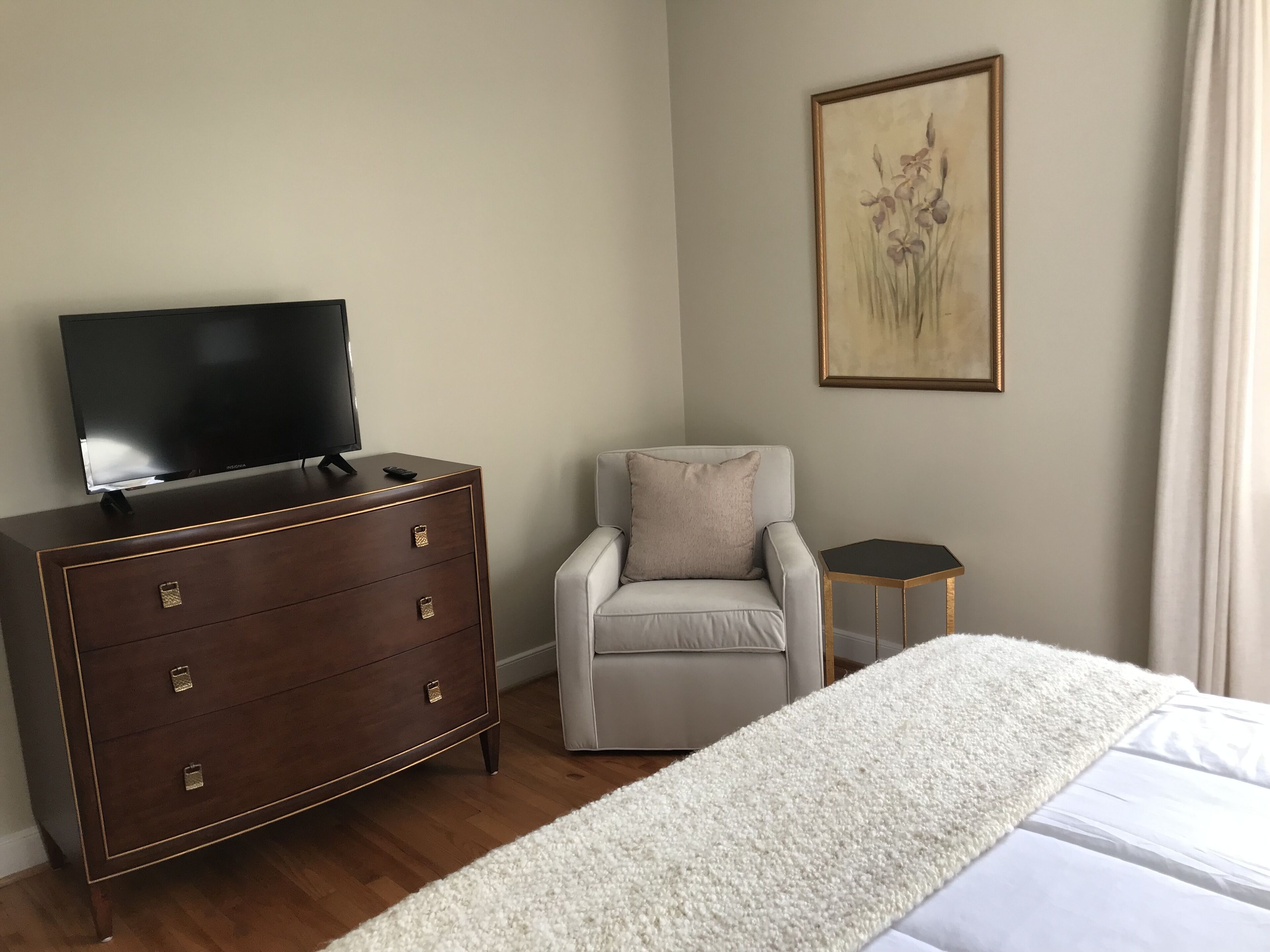 Hotellihuone, jossa on sänky, lipasto, jossa on televisio, tuoli, pieni pöytä ja taulu seinällä.
