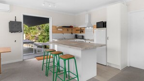 A modern kitchen with white cabinets, a central island with stools, a built-in oven, and a view of greenery outside. - Pearls Embrace - Mangawhai Heads Studio (Mangawhai Heads)