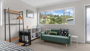 A modern bedroom with a green sofa, a wooden bed frame, a metal wardrobe, and a large window with a view of greenery and buildings. - Pearls Embrace - Mangawhai Heads Studio (Mangawhai Heads)
