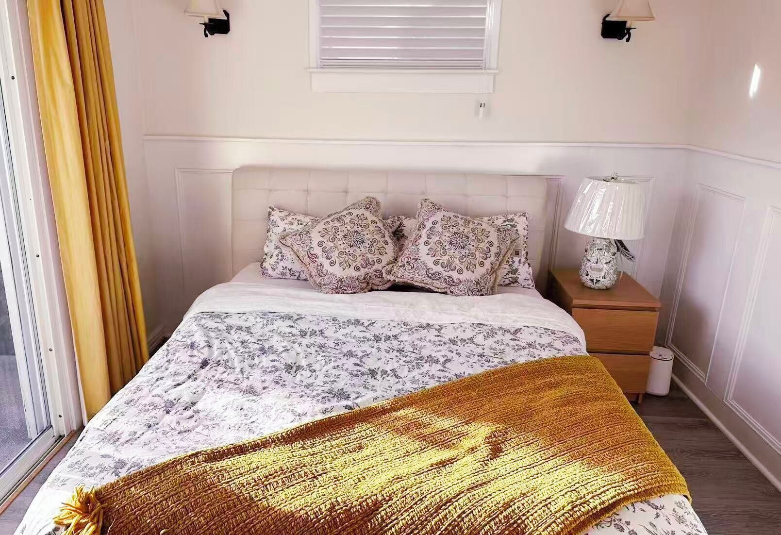 A bedroom with a bed, patterned bedding, a bedside lamp, and a window with blinds.