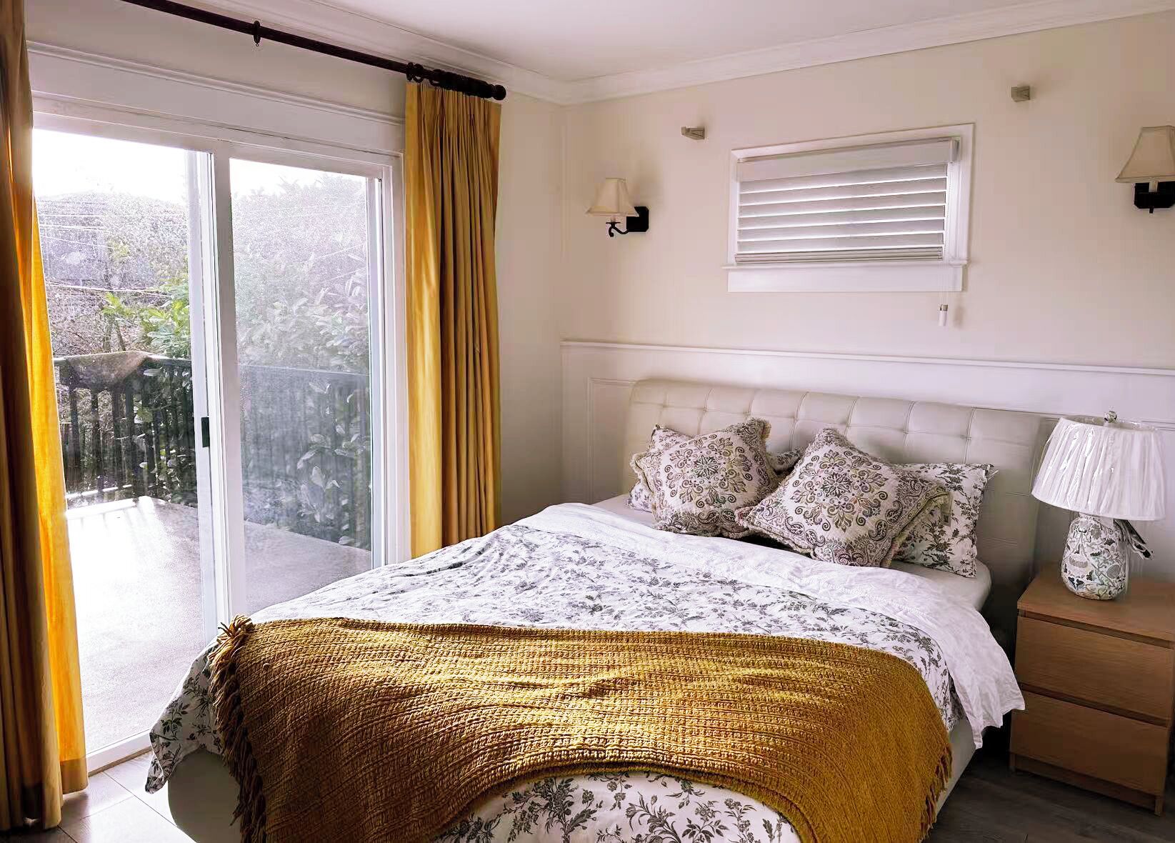 Une chambre à coucher comprenant un lit, une table de chevet, une lampe, une fenêtre avec des stores et une vue sur un balcon.