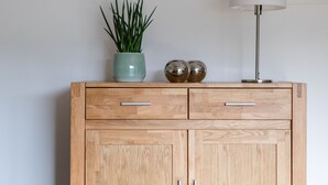 A wooden cabinet with two drawers and two doors, a green plant in a pot, two spherical objects, a white lamp, and a decorative wall hanging. - Ferienhaus am Waldrand Direkt am Sauerländer Höhenflug (Eslohe (Sauerland))