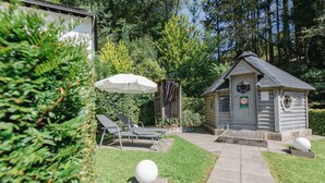 A wooden cabin with a small porch, a covered seating area with chairs and an umbrella, and a stone pathway leading to the cabin. - Ferienhaus am Waldrand Direkt am Sauerländer Höhenflug (Eslohe (Sauerland))