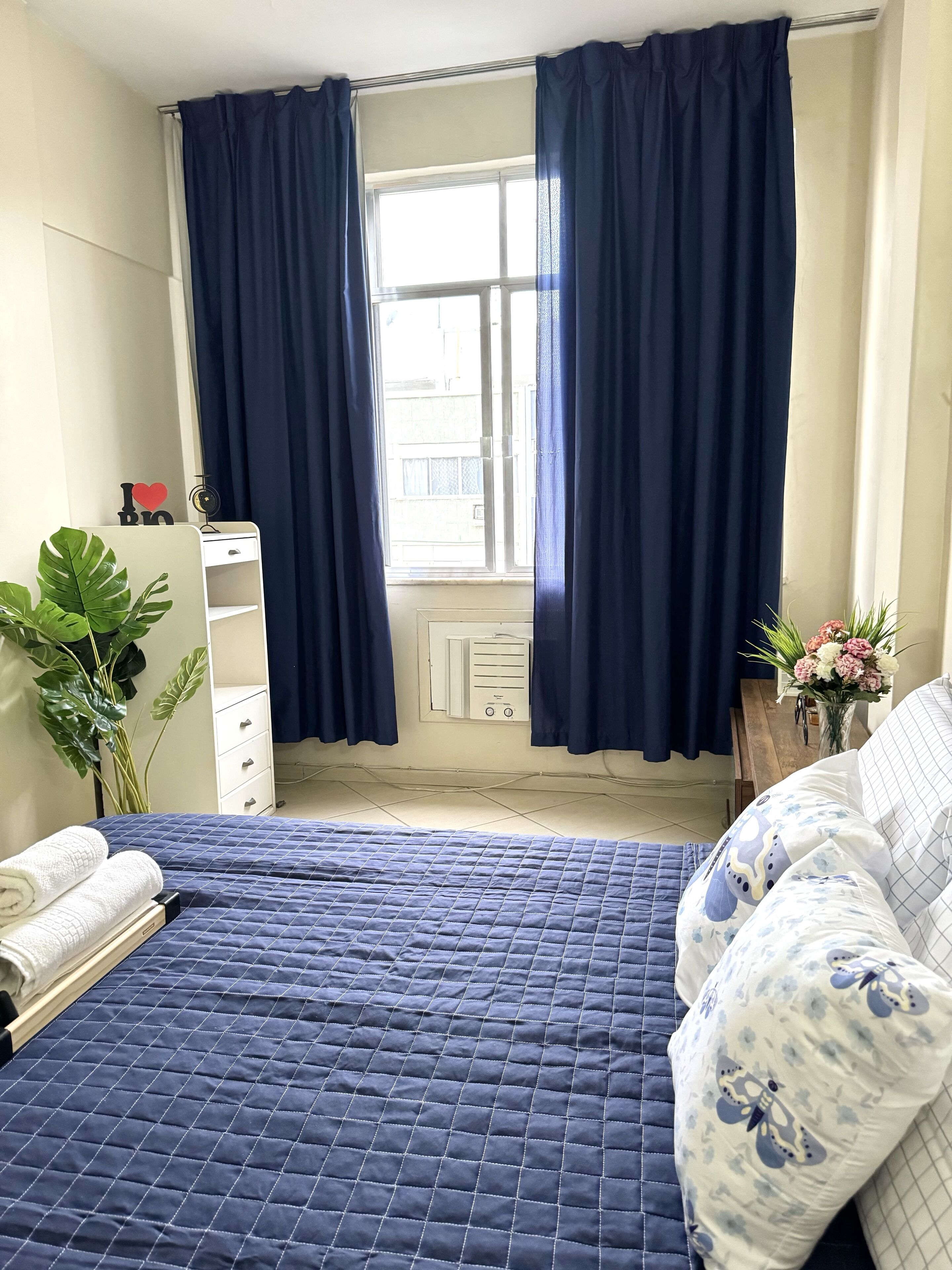 Un dormitorio con una cama, un aparador, un ventanal con cortinas, un radiador y una planta.