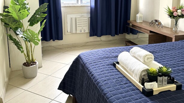 Ein Hotelzimmer mit Bett, einer Zimmerpflanze, einem Nachttisch mit Lampe und einem fenster mit Vorhängen.